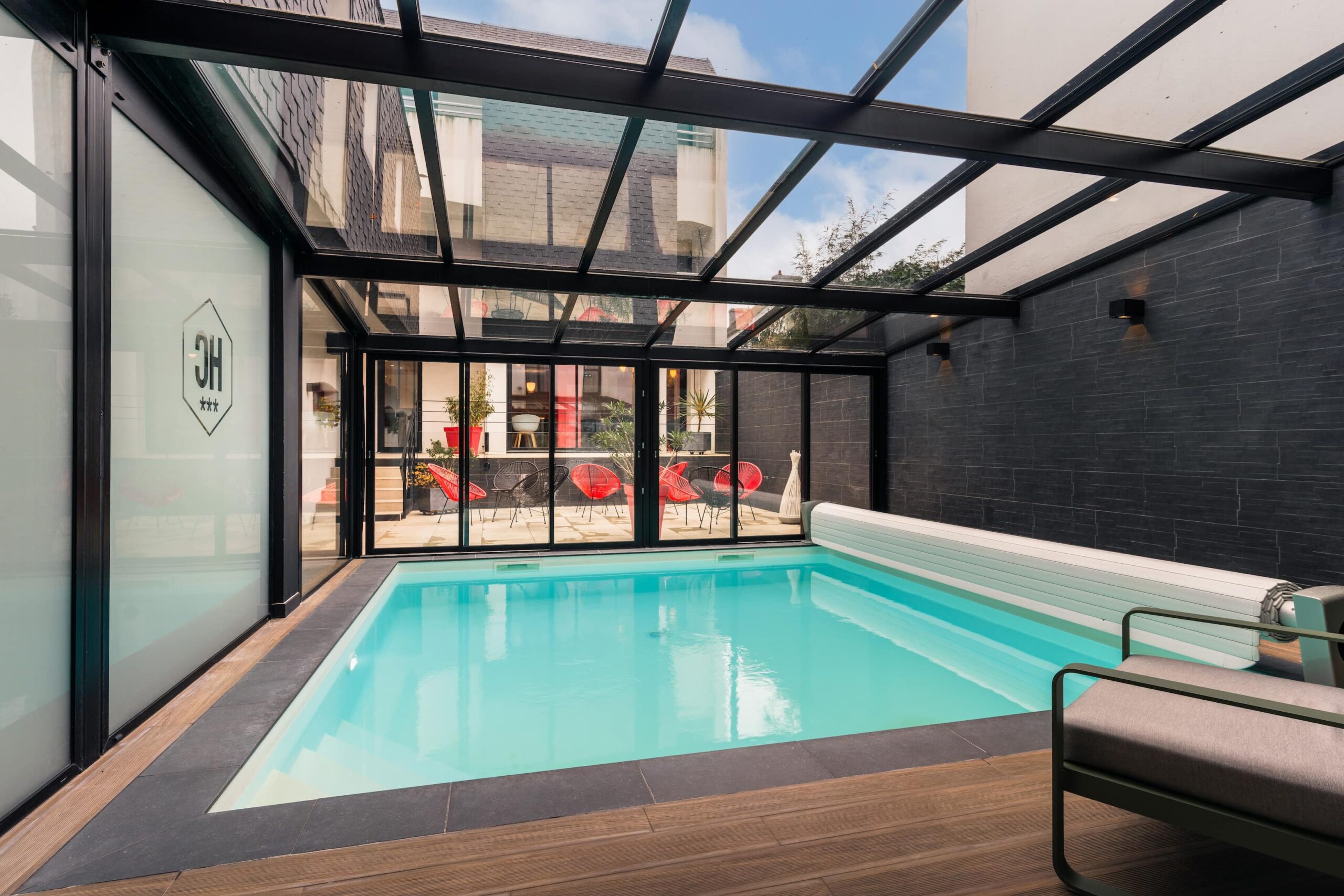 Belle piscine, eau claire, sous verrière et chauffée à l'hôtel LE CORNOUAILLE. Vue sur patio avec siège detente rouge et noir. Design.