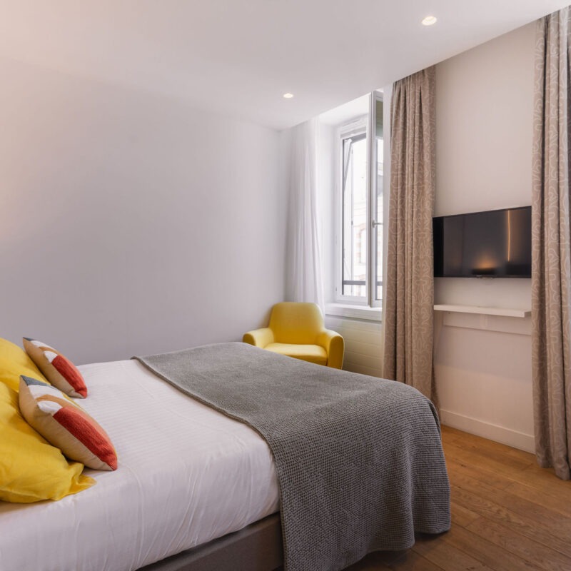 Chambre moderne avec lit double, coussins colorés, fauteuil jaune et télévision murale dans un hôtel à Bénodet.