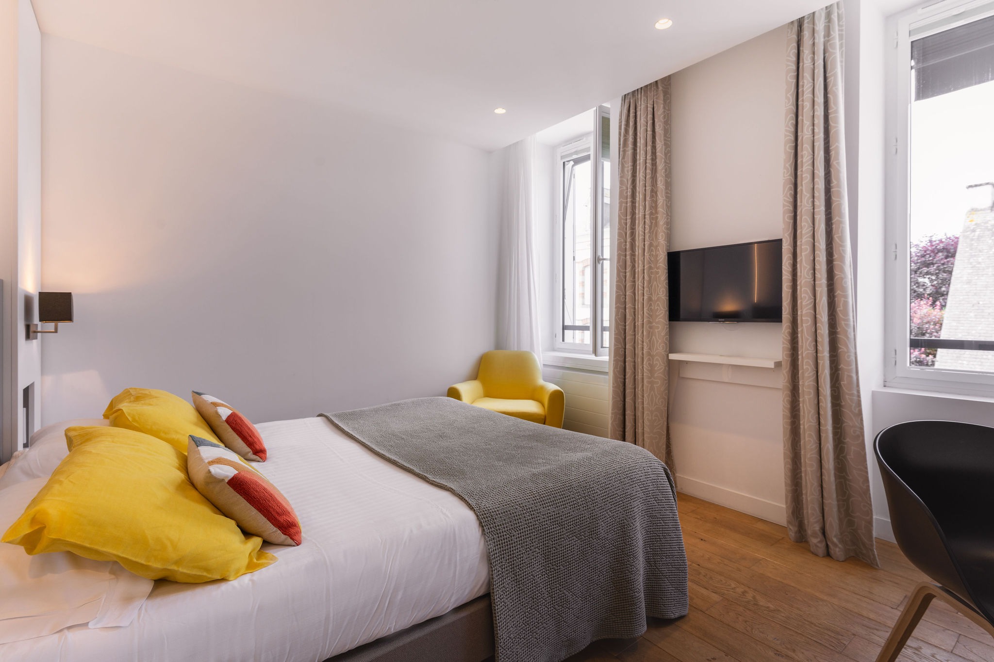 Chambre moderne avec lit double, coussins colorés, fauteuil jaune et télévision murale dans un hôtel à Bénodet.